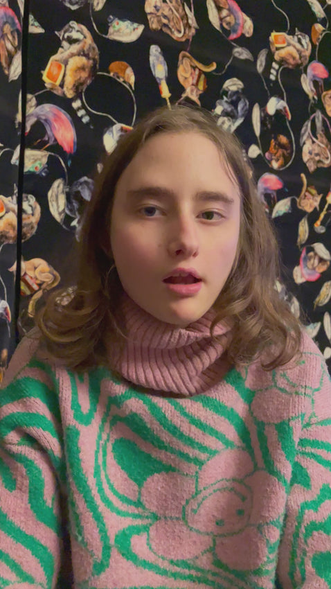 Piper a deaf and autistic teenage artist from hackney reads her note to her mum. she sits in a chair with a pink and green sweater on with a smiley flower on it.