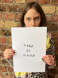 Piper a teenage autistic and deaf artist from hackney hold her Take it easy note in front of her.  The note handwritten in black ink on a white a4 background. Piper wears a leopard print dress and looks to the right over the art and has brick wall behind her.
