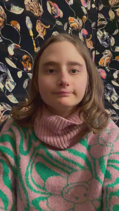Piper a deaf and autistic artist reads her art 'I've believed in you for a long time, my whole life in fact" out excitedly. Piper is sat in a chair with a house of hackney blind behind her and she wears a pink and green knitted jumper with a smiley flower on it.