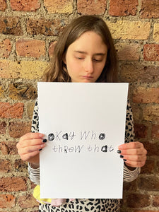 Piper a deaf and autistic teenage artist from hackney is holding her black and white art that she has handwritten in her unique font. she is wearing a leopard print dress and is stood in front of a brick wall. she is looking down at her art.